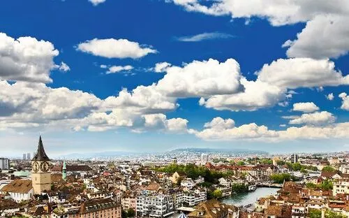 Stadtpanorama mit Fluss und Kirchturm unter blauem Himmel mit Wolken.
