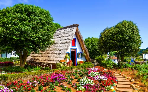 Traditionelles A-förmiges Haus mit Strohdach, umgeben von buntem Garten und Bäumen.