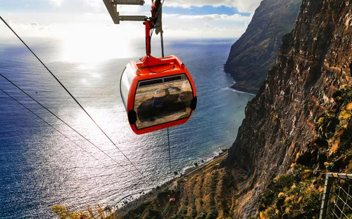 Seilbahnkabine über Küstenschlucht mit Meer im Hintergrund bei Sonnenuntergang.