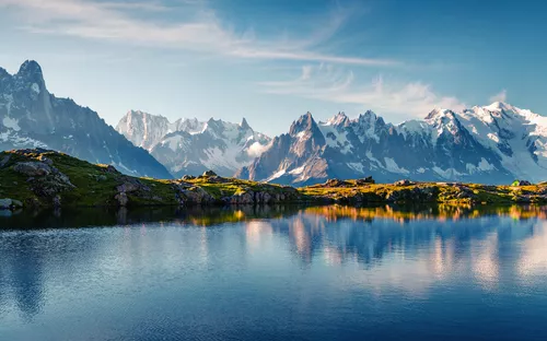 Bergsee mit schneebedeckten Bergen im Hintergrund, ruhiges Wasser, blauer Himmel.