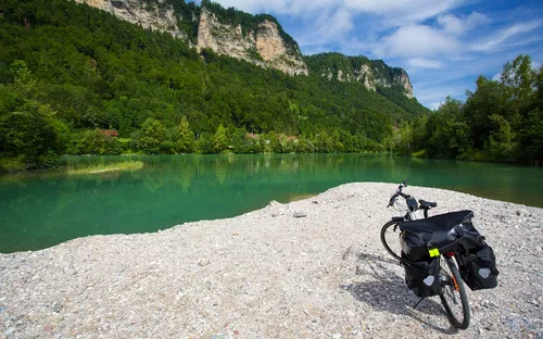 Fahrrad auf Kiesstrand vor grünem See und bewaldeten Bergen bei blauem Himmel.