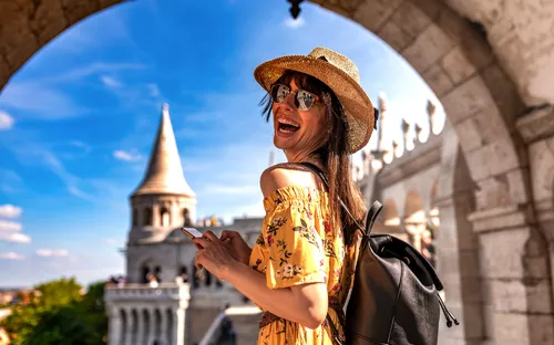 Frau mit Hut und Sonnenbrille lächelt vor historischem Gebäude bei sonnigem Wetter.