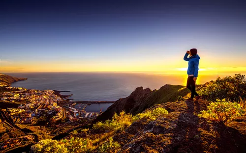 Person auf Berg betrachtet Sonnenuntergang über Stadt und Meer.