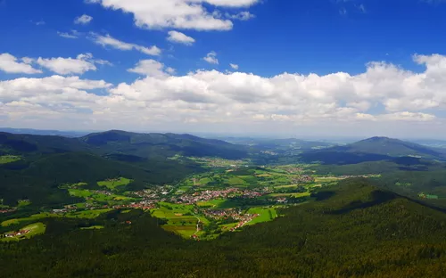 Landschaft mit Hügeln, Wäldern und einer kleinen Siedlung unter blauem Himmel.
