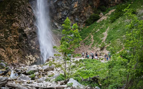 Wasserfall in einer felsigen Landschaft mit grüner Vegetation und Menschen im Hintergrund.