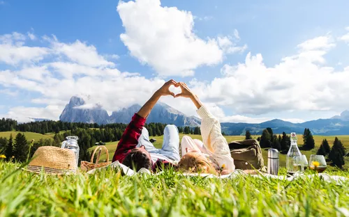 Paar liegt auf Wiese und formt mit Händen ein Herz im Hintergrund einer Berglandschaft.