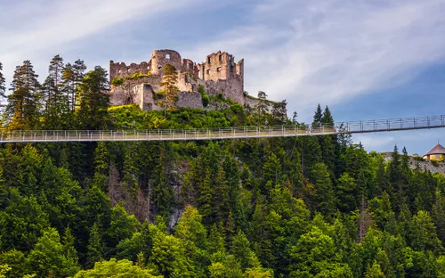 Burg auf einem bewaldeten Hügel mit einer Hängebrücke im Vordergrund.