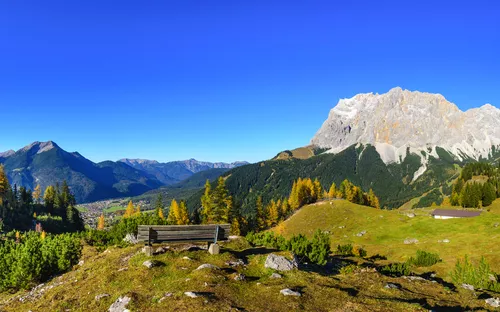 Alpenlandschaft mit Bergen, Wiese und einem Holzbänken unter klarem, blauem Himmel.