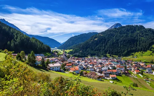 Bergdorf umgeben von grünen Tälern und bewaldeten Bergen unter blauem Himmel.