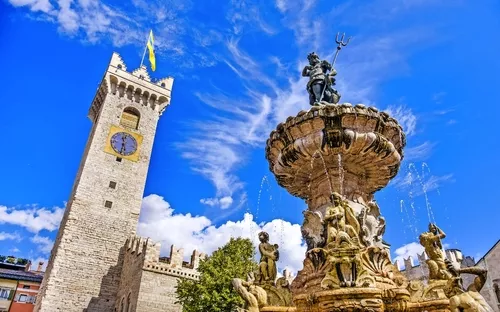 Springbrunnen mit Statuen und Uhrturm im Hintergrund bei blauem Himmel.