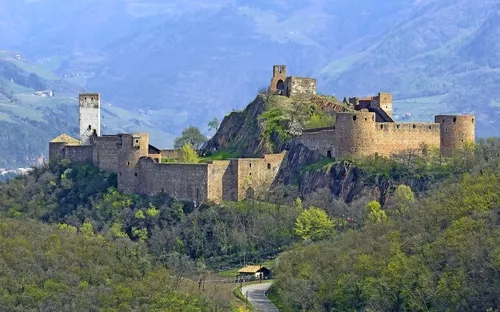 Burganlage auf einem Hügel mit Wald und Bergen im Hintergrund.