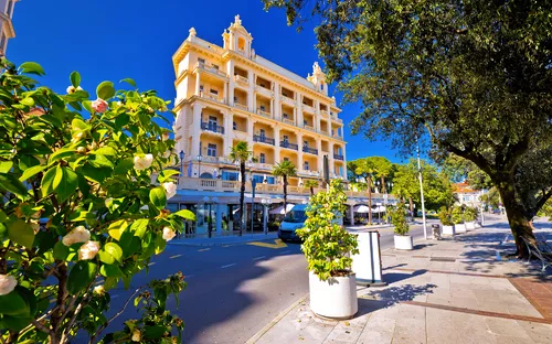 Gelbes Hotel mit Balkonen an einer von Bäumen gesäumten Straße bei blauem Himmel.