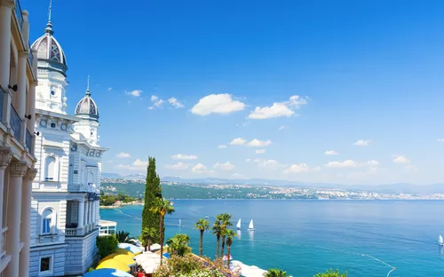 Küstenlandschaft mit historischem Gebäude, Palmen und Segelbooten auf blauem Meer.