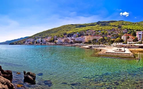 Küstenlandschaft mit klarer Bucht, Häuser im Hintergrund und Statue auf Felsen.