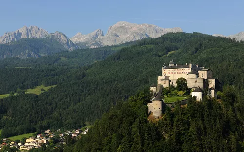Burg auf bewaldetem Hügel, umgeben von Berglandschaft und kleinen Dörfern.