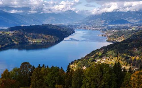 Blick auf einen großen See, umgeben von bewaldeten Hügeln und Bergen im Hintergrund.