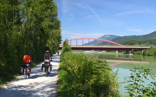 Zwei Radfahrer fahren auf einem Weg entlang eines Flusses, Brücke im Hintergrund.