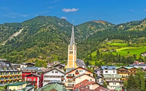 Kirche mit spitzem Turm vor grünen bewaldeten Bergen und bunten Häusern im Vordergrund.