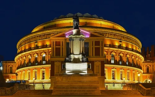 Beleuchtetes, rundes Gebäude bei Nacht mit Statue im Vordergrund.
