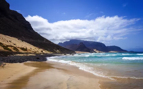 Sandstrand mit Bergen und bewölktem Himmel an der Küste.