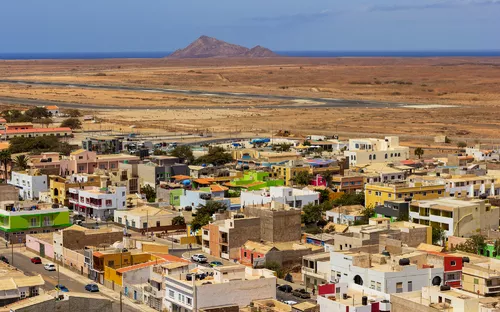 Stadtansicht mit bunten Häusern in einer Wüstenlandschaft, Berg im Hintergrund, blauer Himmel.