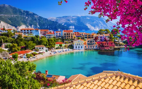 Bunte Häuser am Meer mit blühenden Bäumen im Vordergrund und Bergen im Hintergrund.