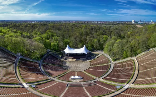 Luftaufnahme eines großen Freilufttheaters mit Wald und Stadt im Hintergrund.