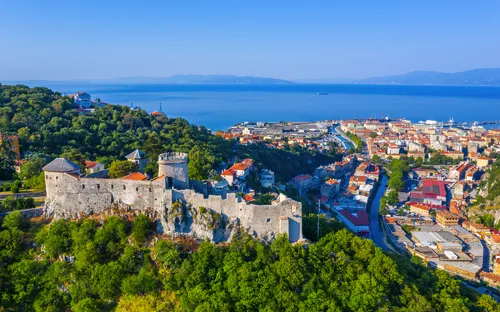 Burg auf Hügel mit Blick auf Küstenstadt und Meer bei klarem Himmel.