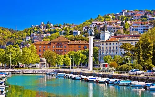 Stadtansicht mit Hafen, Gebäuden und hügeliger grüner Landschaft im Hintergrund.