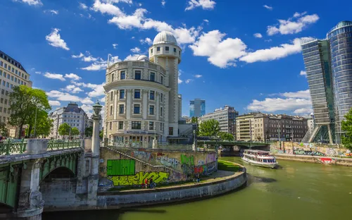 Flussufer mit markantem Gebäude, Brücke und moderner Architektur im Hintergrund.