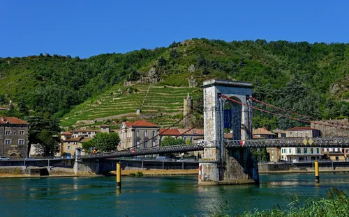 Hängebrücke über Fluss vor hügeliger Landschaft mit Weinbergen und Gebäuden.