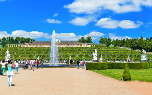 Menschen spazieren im Park vor einer großen Terrassenanlage mit Treppen und Springbrunnen.