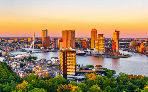 Skyline von Rotterdam bei Sonnenuntergang mit Erasmusbrücke und Hochhäusern am Wasser.