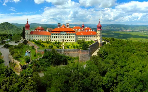 Malerische Burg auf bewaldetem Hügel mit blauem Himmel und Wolken im Hintergrund.