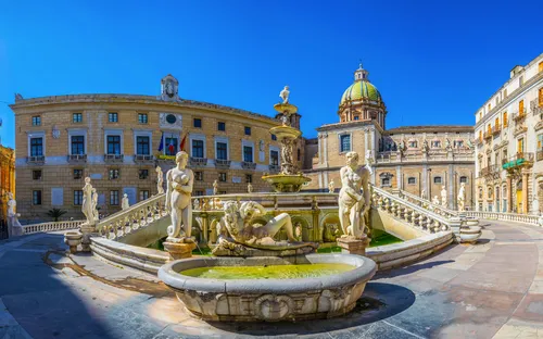 Historischer Brunnen mit Statuen vor alten Gebäuden und blauen Himmel.