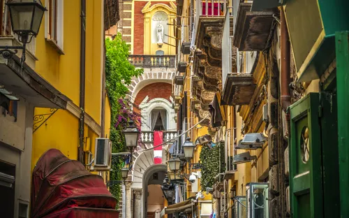 Schmale, belebte Gasse mit gelben Gebäuden und Balkonen in einer historischen Stadt.
