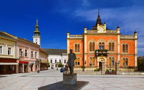 Städtischer Platz mit historischen Gebäuden und einer Statue im Vordergrund.