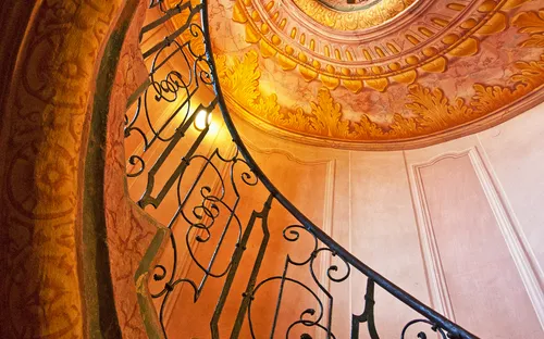 Blick nach oben durch eine verzierte Wendeltreppe mit schmiedeeisernem Geländer.