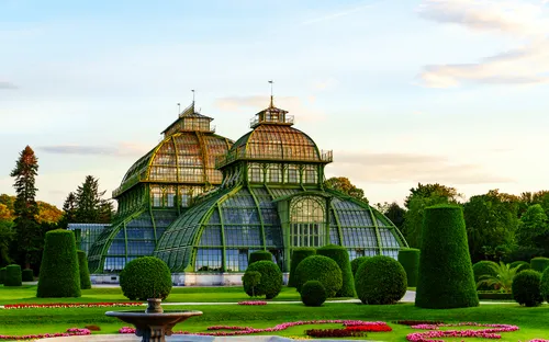 Glaspalast im Garten mit blühenden Beeten und Skulpturen im Hintergrund.
